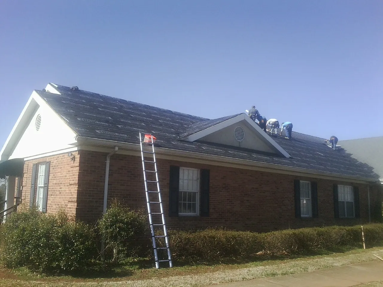 Crew tearing shingles off a brick ranch roof with a ladder set — Emergency Roof Repair work by our crew in Ludington