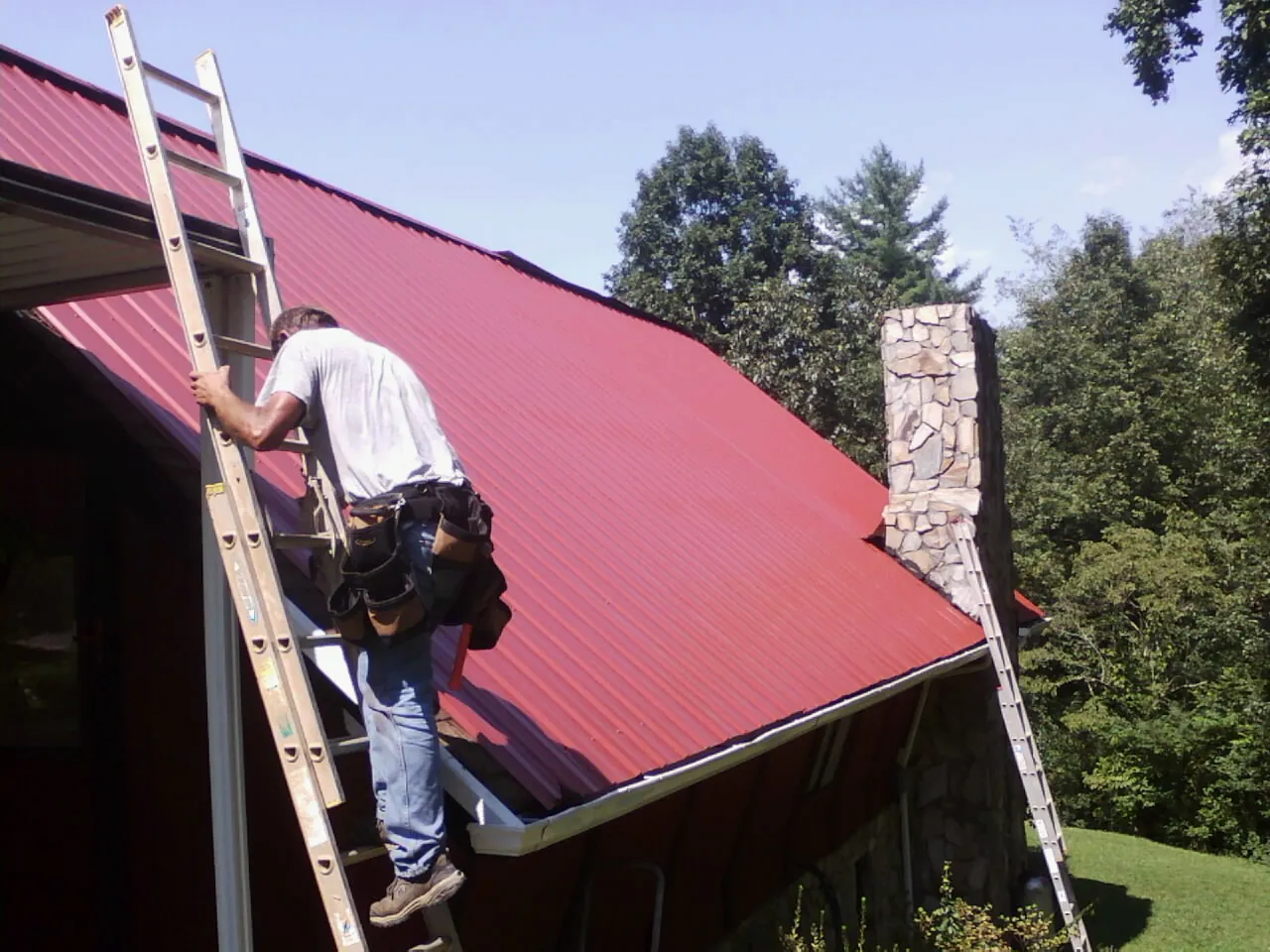 Roofer on a ladder beside a red metal roof and stone chimney — Soffit & Fascia Repair by Roofing Brothers in Ludington